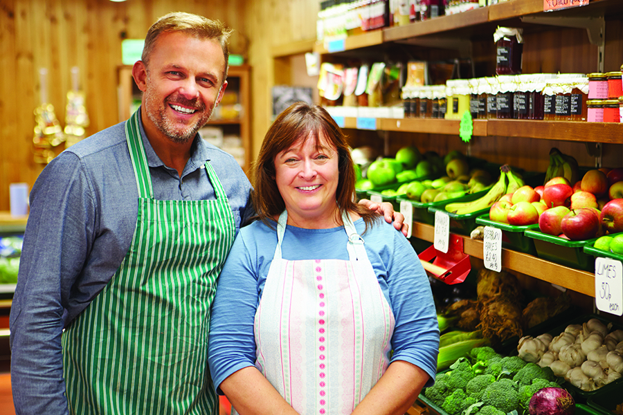 Client Center - Grocery Store Owners Pose in Front of the Produce Section, Wearing Aprons